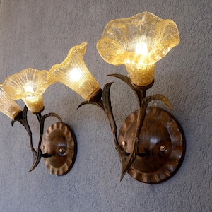May include: Two wall sconces with a floral design. The sconces have amber glass flower shades and bronze metal stems and backplates. They are mounted on a grey wall, creating a warm ambiance. The sconces are decorative lighting.