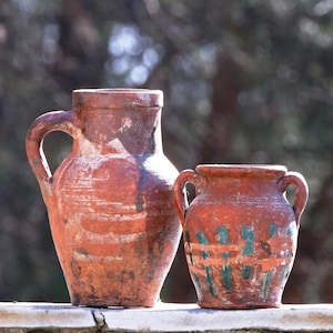 Puede incluir: Dos jarras de cerámica de terracota. La jarra más grande es de color marrón rojizo con un asa. La jarra más pequeña tiene dos asas y detalles pintados en verde y blanco. Aspecto rústico.