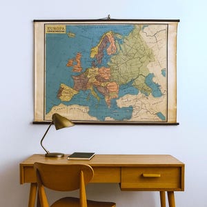Könnte beinhalten: Eine Vintage-Karte von Europa mit der Aufschrift "EUROPA" hängt an einer Wand. Die Karte ist in Blau-, Grün- und Brauntönen gehalten. Darunter befindet sich ein Holzschreibtisch mit einer kleinen Lampe und einem Notizbuch. Ein Holzstuhl steht vor dem Schreibtisch.