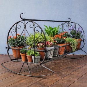 May include: A black wrought iron plant stand with a rocking base, holding several terracotta pots filled with various green and orange flowering plants. The stand has decorative scrollwork and is set on a wooden deck.
