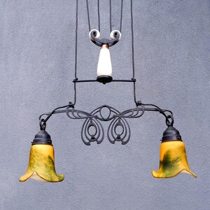 May include: An antique-style chandelier with a black wrought iron frame. It features two bell-shaped lampshades with yellow and green glass. The fixture has a decorative design and a white and brown central element.