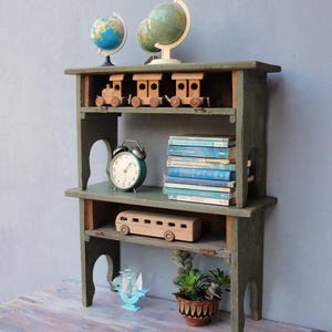 May include: A distressed green wooden shelf unit with three levels. The top shelf holds two globes and three wooden toy trains. The middle shelf has a green alarm clock and a stack of blue books. A wooden bus and a small plant are on the bottom shelf.