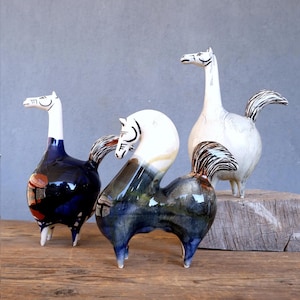 Puede incluir: Tres esculturas de caballos de cerámica con cuellos largos y rasgos estilizados. Los caballos tienen un acabado brillante en tonos azules, blancos y marrones. Se alzan sobre una superficie de madera sobre un fondo gris. Piezas de arte decorativas.