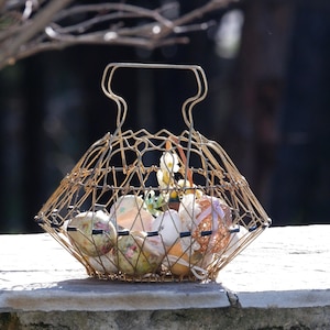 May include: A gold wire basket, filled with decorated Easter eggs. The eggs feature floral and glitter designs. The basket has a handle and sits on a stone wall, with a blurred background of branches and dark foliage.