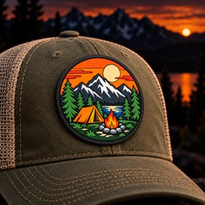 May include: An olive green baseball cap with a mesh back and a circular patch on the front. The patch features a colourful graphic of a tent, a campfire, pine trees, a lake, and snow-capped mountains under a bright orange sunset sky.
