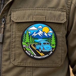 May include: A circular patch featuring a light blue car driving on a winding road through a forest with snow-capped mountains and a yellow sun in the background. The text MADE BY PROMOTIONAL LAB is printed along the bottom edge of the patch.