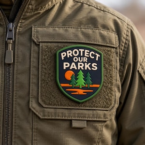 May include: A shield-shaped patch attached to an olive green jacket. The patch features a dark blue background with the text PROTECT OUR PARKS in cream letters, a bright orange sun, three green pine trees, and an orange landscape.