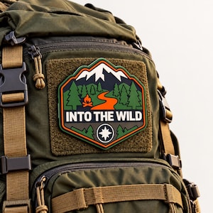 May include: An olive green tactical backpack featuring a rectangular hook-and-loop patch. The patch displays a mountain landscape with pine trees, a campfire, a winding orange path, and the text INTO THE WILD above a compass star icon.