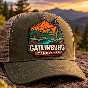 May include: An olive green trucker cap featuring a patch with a mountain landscape, a cabin, a cable car, and a river. The patch includes the text GATLINBURG TENNESSEE in bold lettering. The cap has a mesh back and a structured front panel.