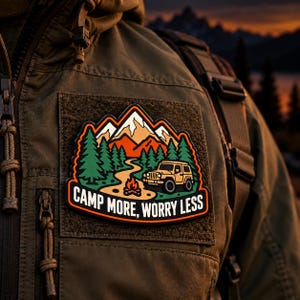 May include: A rectangular patch attached to an olive green jacket featuring a graphic of a yellow off-road vehicle, a campfire, green pine trees, and orange mountains. The text "CAMP MORE, WORRY LESS" is printed in white at the bottom.