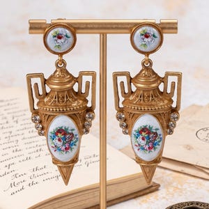May include: A pair of gold-toned drop earrings shaped like miniature urns, featuring white enamel oval insets with colorful floral patterns, small clear crystals along the sides, and round floral studs, displayed on a gold metal T-bar stand.