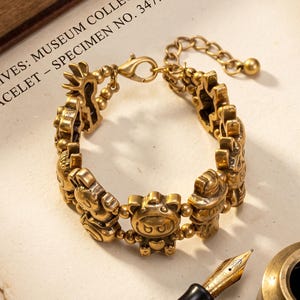 Può includere: Un bracciale con ciondoli in metallo color oro caratterizzato da una serie di piccole figure scolpite collegate da perline sferiche. Il bracciale include una chiusura a moschettone e una catenella di estensione. Testo: ARCHIVES: MUSEUM COLLECTION BRACELET - SPECIMEN NO. 347.