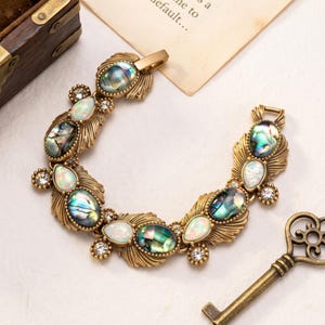 May include: A gold-toned metal bracelet featuring oval abalone shell cabochons and white iridescent teardrop stones. The design includes small clear rhinestones and textured leaf-like metal settings arranged in a curved, ornate link pattern.