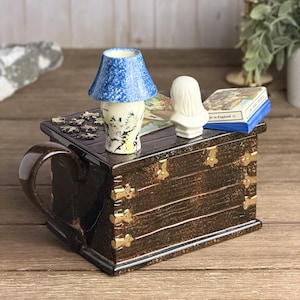May include: A dark brown ceramic teapot shaped like a wooden trunk with gold-coloured metal accents. The lid features a miniature blue speckled lamp, a white bust figurine, and a small box labelled JIG-SAW PUZZLE Made in England on the top surface.