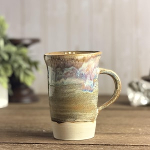 May include: A tall ceramic mug featuring a cylindrical body with a handle on the right side. The base is a solid matte cream colour, transitioning upwards into a textured, earthy brown midsection, topped with a glossy, dripped glaze of blue and purple.