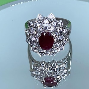 May include: A silver-toned ring featuring a central oval-cut deep red gemstone surrounded by two concentric layers of small, clear, sparkling stones arranged in a starburst pattern, set on a polished metal band.