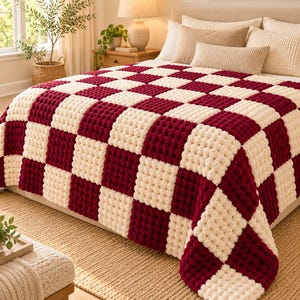 May include: A chunky crochet blanket featuring a repeating checkerboard pattern in deep burgundy and cream white. The blanket is constructed from thick, textured yarn squares arranged in a grid, draped over a bed with neutral-toned pillows.
