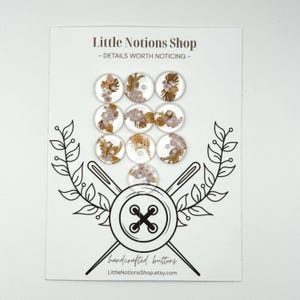 May include: Ten circular buttons arranged on a white display card labeled Little Notions Shop. Each button features a clear surface with embedded gold-toned botanical leaf and small pink floral designs. The card displays a black line drawing of a button and needles.