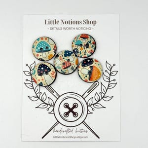 May include: Five round buttons featuring colorful mushroom illustrations on a white display card. The card displays the text Little Notions Shop - DETAILS WORTH NOTICING - and a graphic of two crossed needles over a large button with the text handcrafted buttons.