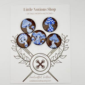 May include: Five round brown buttons with two holes each, featuring blue and white hand-painted mushroom illustrations. The buttons are displayed on a white card labeled Little Notions Shop with a graphic of crossed needles and a button.