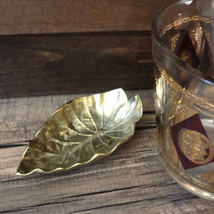 May include: A gold leaf-shaped dish with a textured surface sits on a wooden table next to a glass jar with a gold and burgundy design. The jar contains a set of gold tongs.