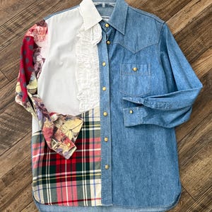 May include: A patchwork shirt featuring denim, white, floral, and plaid patterns. The shirt has a collared neckline, long sleeves, and gold-colored button closures. The left side of the shirt has a white panel with a ruffled trim.