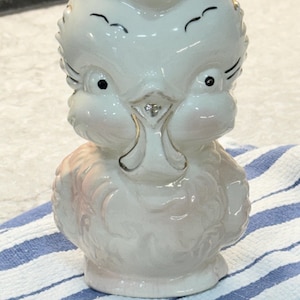 May include: A white ceramic owl figurine with pink and gold accents. The owl has large, black eyes and a wide beak. The figurine is sitting on a blue and white striped surface.