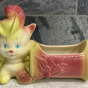 May include: A vintage ceramic planter shaped like a cat wearing a hat. The planter is painted in shades of pink, yellow, and green. The cat has blue eyes and a pink nose. The planter has a rectangular opening for planting.