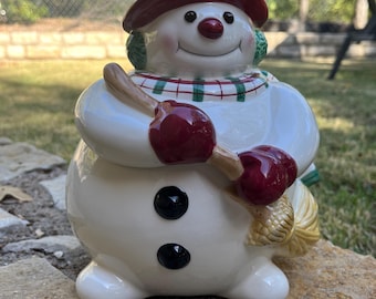 Peut inclure: Figurine de bonhomme de neige en céramique avec un chapeau, des cache-oreilles et des mitaines bordeaux. Le bonhomme de neige a un nez en carotte, des yeux en bouton noirs et un visage souriant. Il tient un balai et porte une écharpe à carreaux verts et rouges.