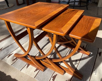 Puede incluir: Un conjunto de tres mesas nido de madera con un acabado en madera natural. Las mesas tienen un diseño curvo único y están apiladas una encima de la otra.