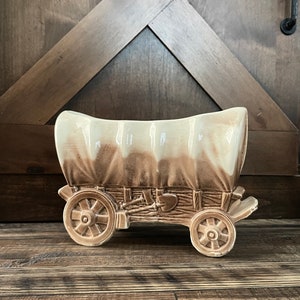 May include: A ceramic planter shaped like a covered wagon. The wagon is painted in shades of brown and cream. The planter is a great way to add a rustic touch to your home decor.