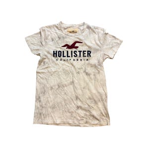 Puede incluir: Una camiseta de manga corta con un patrón tie-dye en tonos blanco roto y gris. El pecho presenta un logotipo de gaviota rojo oscuro sobre el texto azul marino Hollister California. Una etiqueta de marca es visible en el cuello.