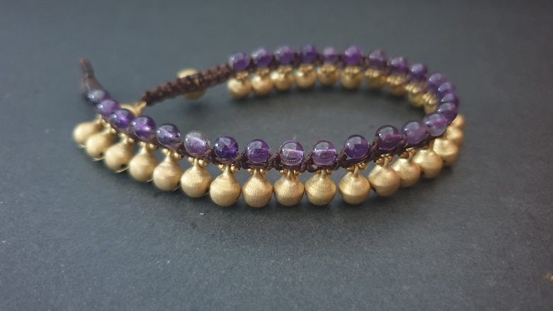 May include: A purple amethyst and gold bead bracelet with a brown cord. The bracelet has a double row of beads with gold bells hanging from the bottom row.