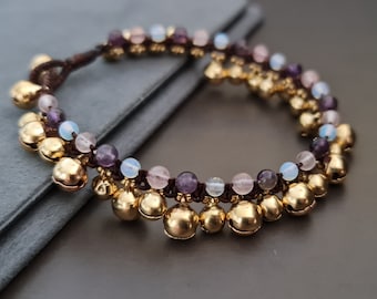 Pode incluir: Um bracelete de contas multicoloridas com sinos dourados. As contas são em tons de roxo, rosa e azul.