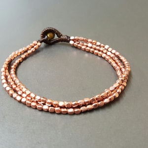 May include: A copper-colored bracelet with three strands of small, square beads. The bracelet is tied with a brown cord and has a knot closure. The beads have a metallic sheen, and the bracelet is displayed on a gray surface.