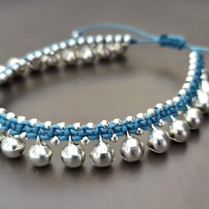 May include: A blue and silver ankle bracelet with small bells. The bracelet is made of braided cord and has a silver clasp.