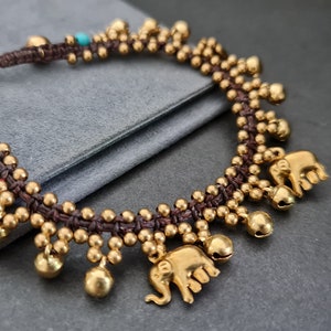 May include: A brown cord bracelet with gold beads and bells, featuring two small elephant charms. The bracelet has turquoise beads interspersed throughout.