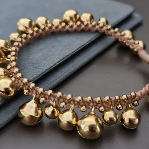 Könnte beinhalten: Ein rosafarbenes Armband aus Schnur mit goldenen Glöckchen. Das Armband besteht aus einer dünnen, geflochtenen Schnur und hat eine Reihe kleiner, runder Glöckchen, die daran befestigt sind.