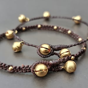 Puede incluir: Dos pulseras trenzadas marrones con cascabeles dorados. Las pulseras están atadas juntas y tienen varios cascabeles dorados.
