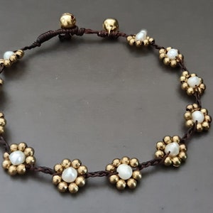 Op de afbeelding: Een bruin koordarmband met gouden en witte kralen in de vorm van bloemen.