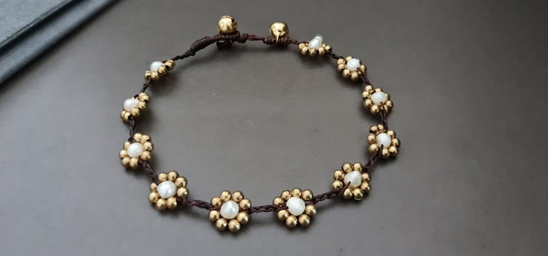 Op de afbeelding: Een bruin koordarmband met gouden en witte kralen in de vorm van bloemen.