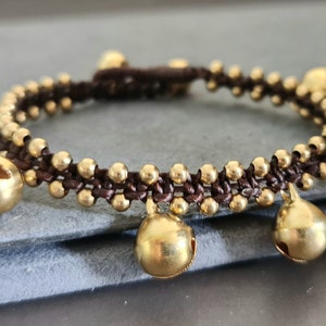Könnte beinhalten: Ein braunes geflochtenes Armband mit goldfarbenen Glöckchen, die daran hängen. Das Armband liegt auf einer grauen Oberfläche.