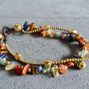 May include: A gold bracelet with a brown cord and colourful gemstone beads. The bracelet has small gold bells attached.
