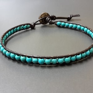 Puede incluir: Un brazalete de cuero marrón con cuentas turquesas. El brazalete tiene un cierre simple de nudo.