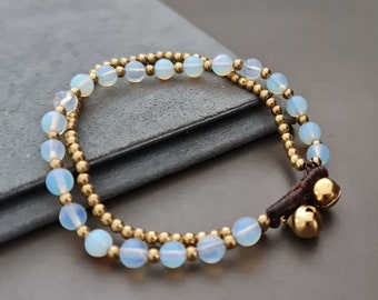 May include: A double-strand bracelet with white opal beads and gold accents. The bracelet has two small bells attached to a brown leather cord.