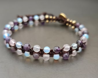 Pode incluir: Um bracelete de várias voltas com contas de ametista, quartzo rosa e pedra da lua. O bracelete é feito com cordão castanho e tem um fecho dourado.