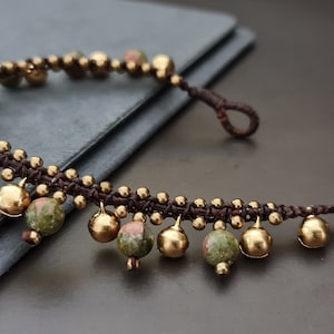 May include: A brown cord bracelet with gold and green gemstone beads. The bracelet has a dangling design with multiple beads hanging from the cord.