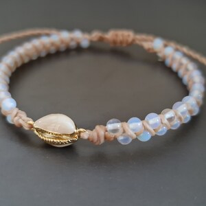 May include: A handmade bracelet featuring a natural seashell pendant with gold-colored trim. The bracelet is made with beige macrame cord and iridescent beads. The adjustable design allows for a comfortable fit.