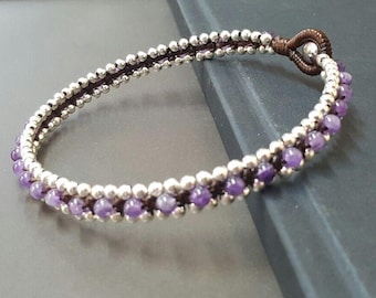 Pode incluir: Um bracelete de pedra preciosa de ametista roxa com contas prateadas e cordão marrom.