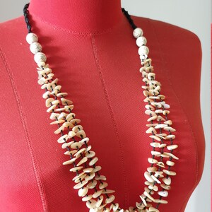 Op de afbeelding: Een meerstrengs ketting gemaakt van witte en beige schelp kralen met zwarte koord accenten. De ketting wordt getoond op een rood manekin.